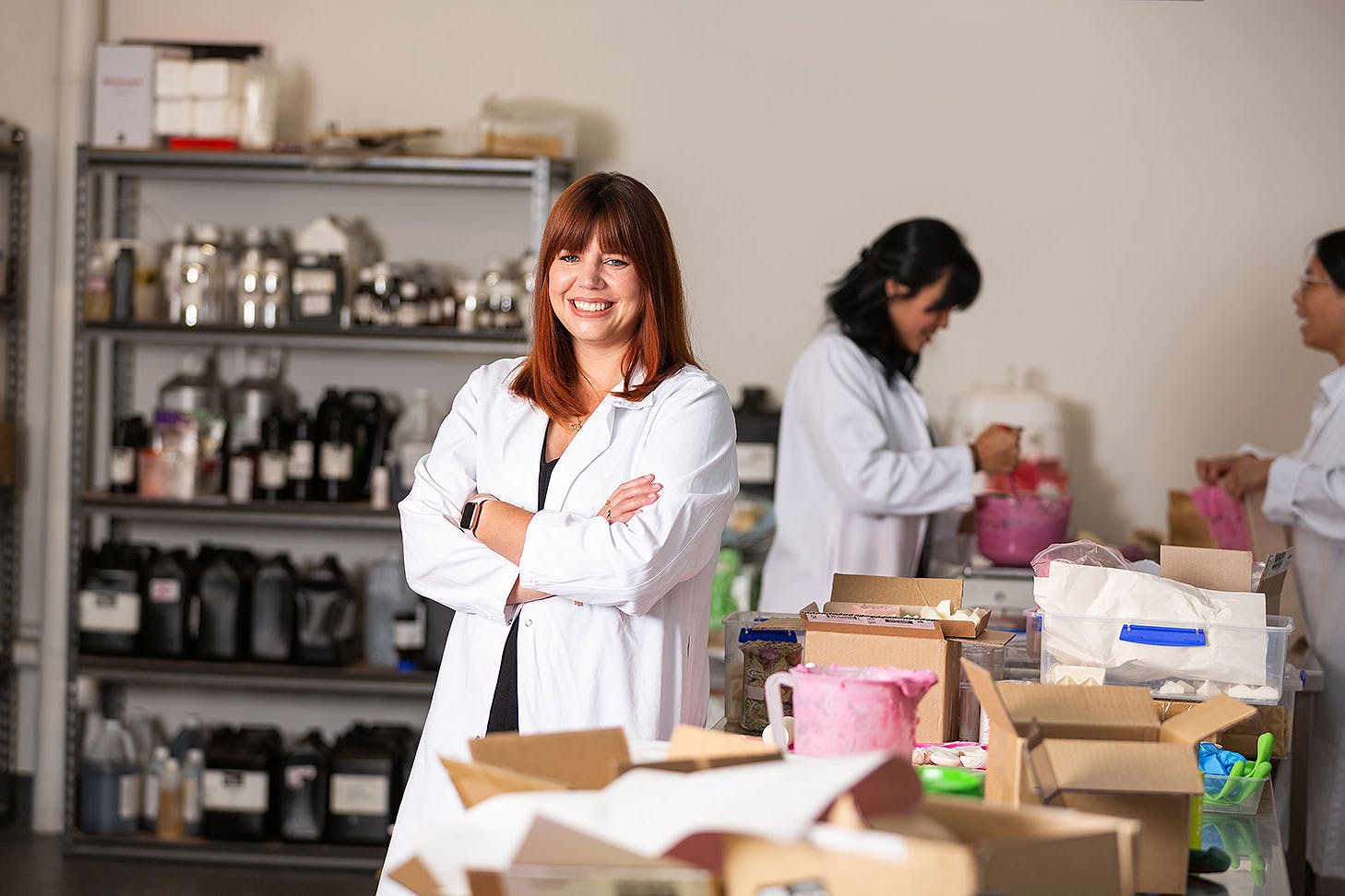 Brianne wearing a white lab coat in Ethique's manufacturing space, with two women making soap mix in the background.